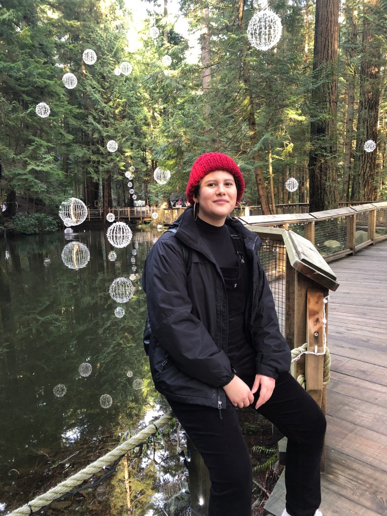 Maria with short hair and a red beanie in an all black outfit sitting on a rail next to a pong in the Capilano Bridge Park in Vancouver, Canada 